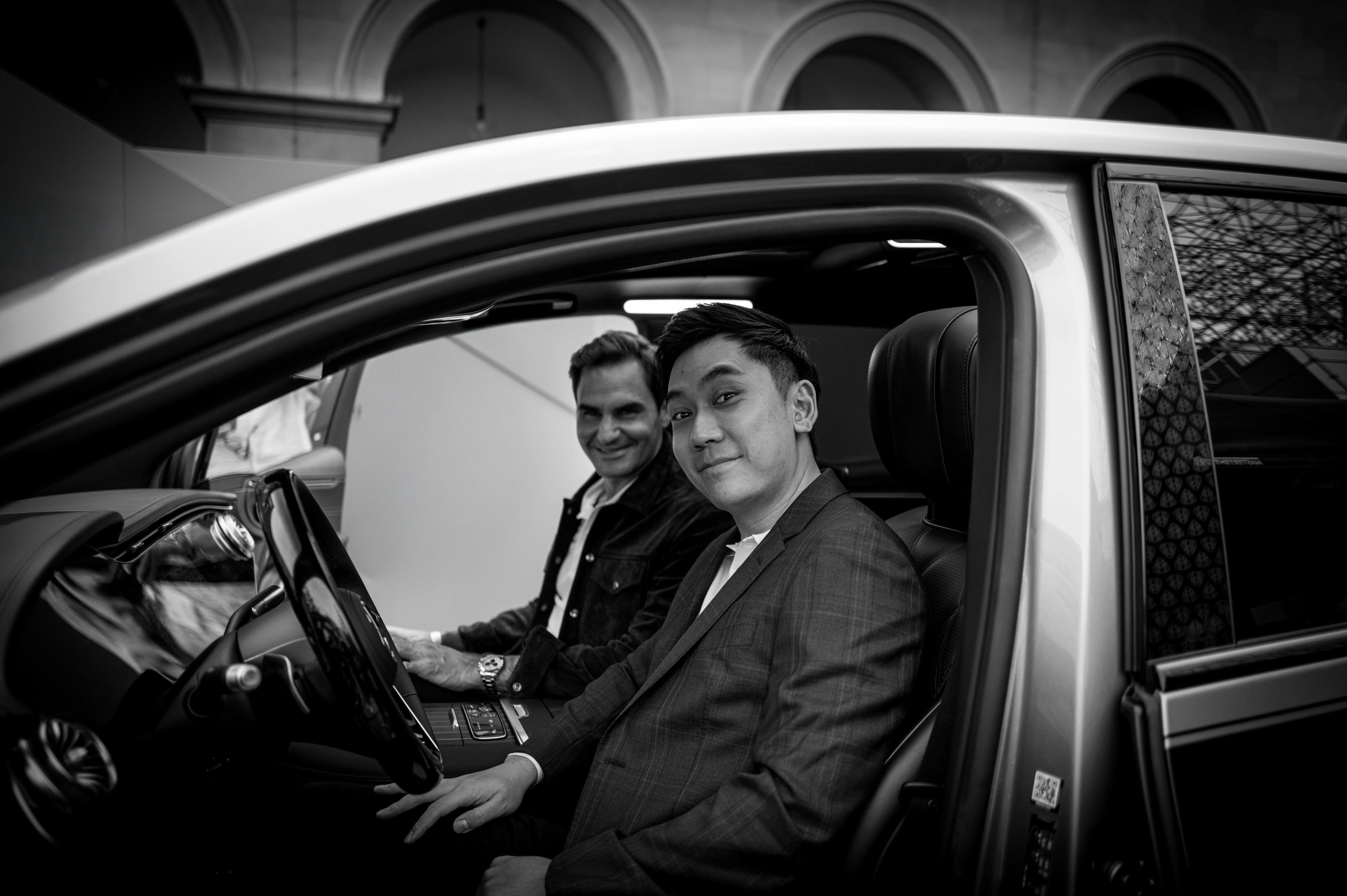 Two men (Peter Yan and Roger Federer) sitting inside a car, one driving and the other sitting in the passenger seat.